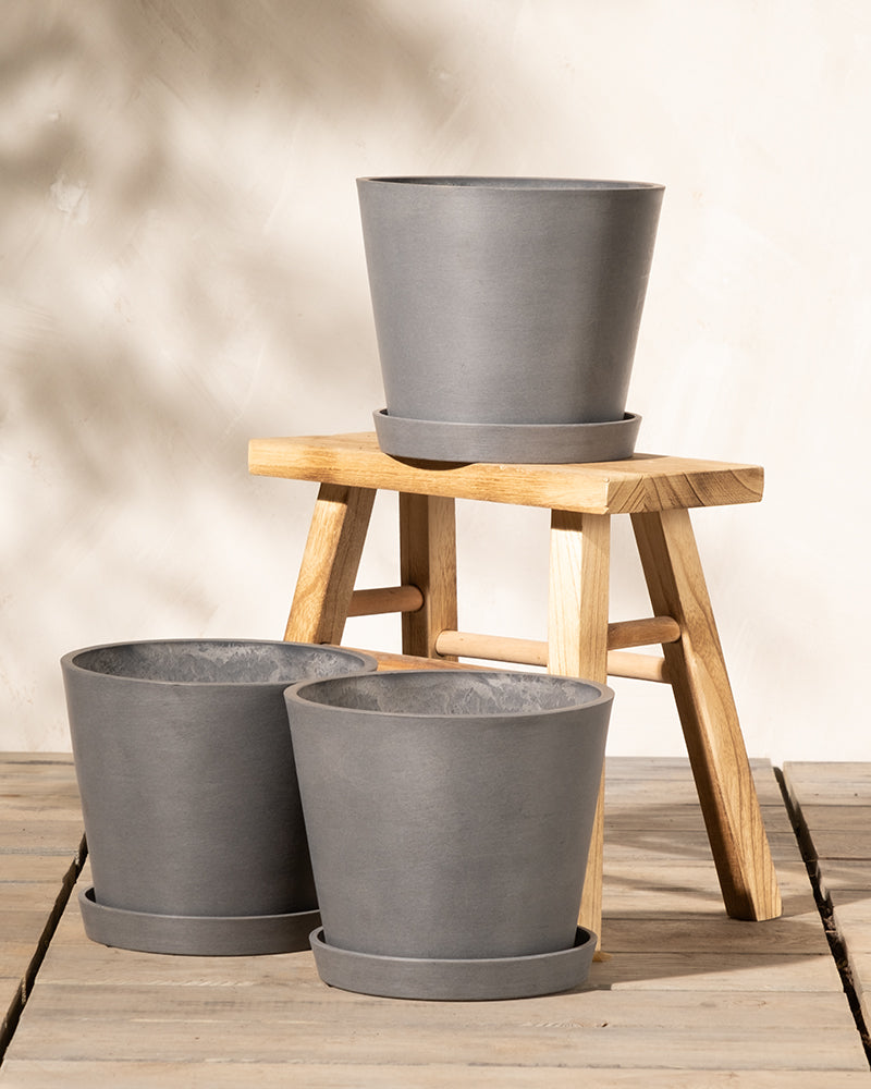 Ausgestellt sind drei graue Pflanztöpfe aus Keramik, zwei davon auf einer Holzfläche und einer auf einer kleinen Holzbank. Unter jedem Pflanzkübel-Set Amsterdam (3x 20) steht ein passender Untersetzer. Der Hintergrund ist eine neutrale, helle Wand. Die Szene ist hell und minimalistisch.