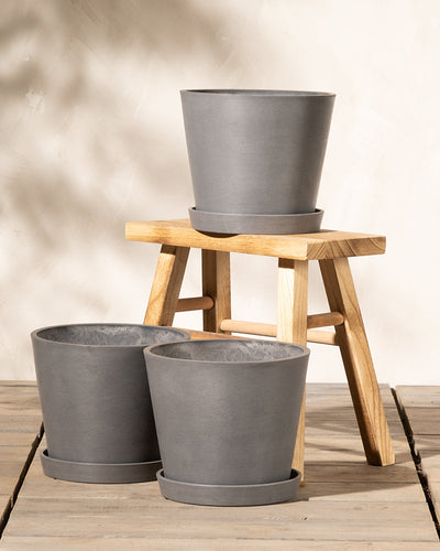 Ausgestellt sind drei graue Pflanztöpfe aus Keramik, zwei davon auf einer Holzfläche und einer auf einer kleinen Holzbank. Unter jedem Pflanzkübel-Set Amsterdam (3x 20) steht ein passender Untersetzer. Der Hintergrund ist eine neutrale, helle Wand. Die Szene ist hell und minimalistisch.