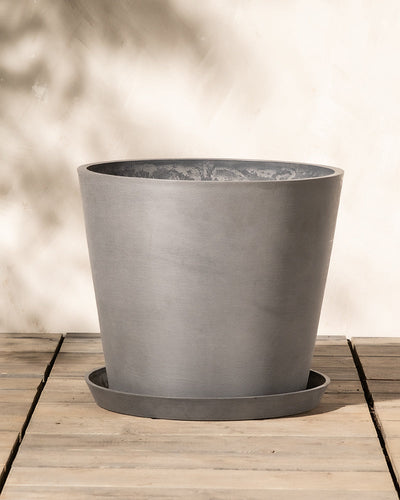 Ein großer, runder Blumentopf aus grauer Keramik auf einer Holzoberfläche im Freien. Der Kunststoff-Pflanzkübel (Amsterdam | 40 cm ⌀) hat einen passenden Untersetzer an seiner Unterseite und verfügt über ein effizientes Entwässerungssystem. Vor einem beigen Hintergrund mit weichem, natürlichem Licht, das schwache Schatten wirft.