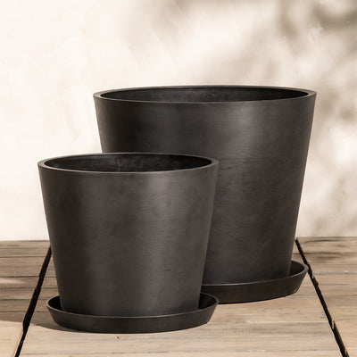 Zwei schwarze, minimalistische Töpfe des Pflanzkübel-Sets Amsterdam (40, 30) mit matter Oberfläche stehen auf einer Holzfläche vor einer hellen Wand. Ein Topf ist größer und etwas hinter dem kleineren platziert. Beide Töpfe stehen auf passenden runden Tabletts mit einem effizienten Drainagesystem und werfen sanfte Schatten an die Wand.