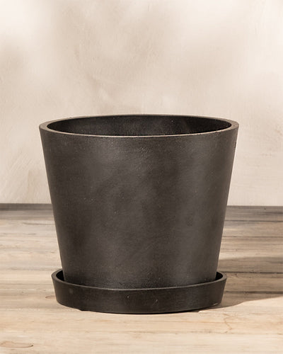 Ein einfacher schwarzer Kunststoff-Pflanzkübel (Amsterdam | 20 cm ⌀) von EcoPots mit passendem Untersetzer steht auf einer Holzoberfläche vor einem hellen, strukturierten Hintergrund. Der aus Recycling-Kunststoff gefertigte Topf hat eine breite Öffnung, die sich unten verjüngt, und der Untersetzer passt genau darunter, um überschüssiges Wasser aufzufangen.