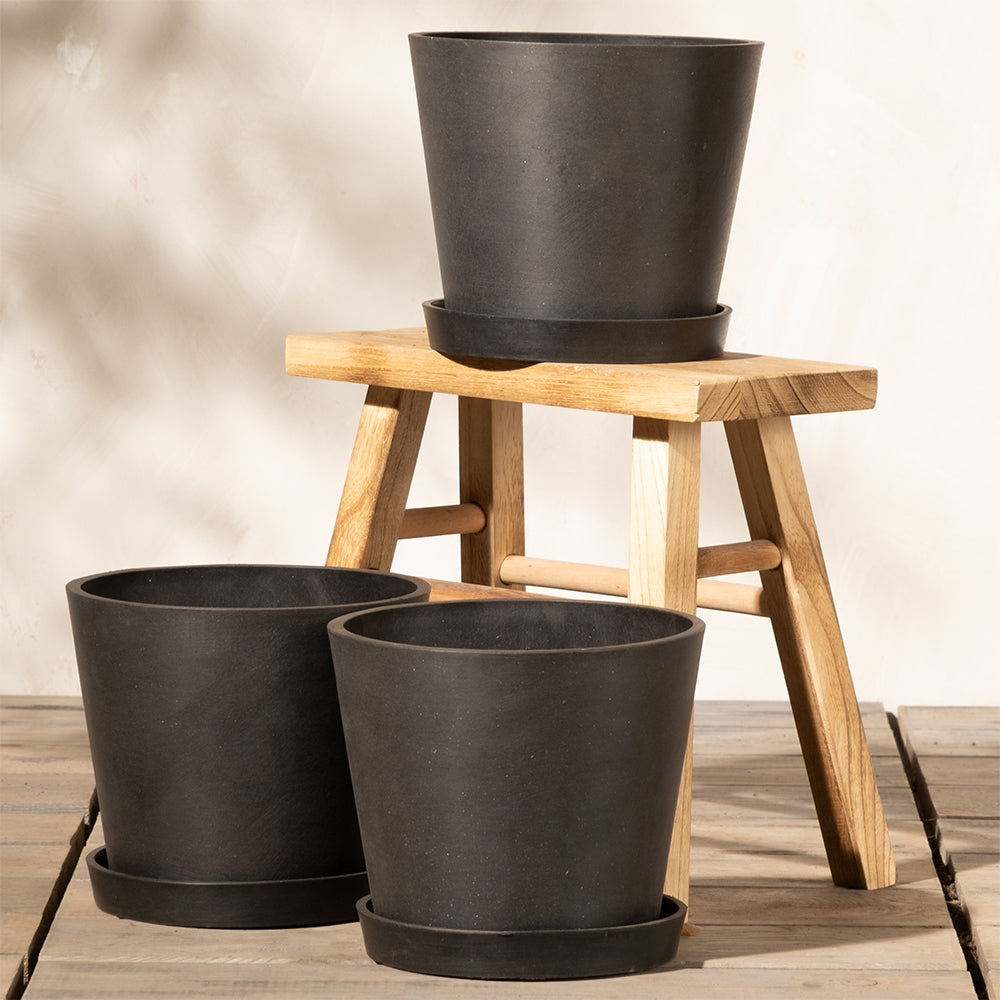 Drei schwarze Pflanzkübel-Set Amsterdam (3x 20) mit Untersetzern stehen auf einem Holzhocker auf einer Holzfläche. Zwei stehen auf dem Boden, während einer auf dem Hocker sitzt und sein effizientes Entwässerungssystem zur Schau stellt. Der Hintergrund ist eine schlichte, helle Wand.