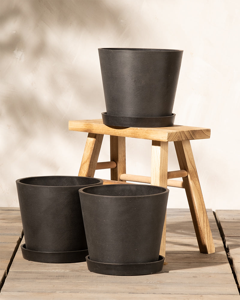 Ein minimalistisches Indoor-Setup mit drei schwarzen Pflanzkübel-Set Amsterdam (3x 20) mit Untersetzern. Zwei Töpfe stehen auf einer Holzfläche, während der dritte Topf erhöht auf einem kleinen Holzhocker steht. Der Hintergrund ist eine rustikale, helle Wand. In den Töpfen sind keine Pflanzen zu sehen, die über ein effizientes Drainagesystem verfügen.