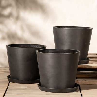 Drei schwarze, zylindrische Pflanzkübel-Set Amsterdam (3x 30) in verschiedenen Größen stehen auf einer Holzoberfläche vor einer neutralen Wand. Jeder Topf besteht aus Recycling-Kunststoff und steht auf einem passenden Untersetzer mit einem effizienten Abflusssystem, das überschüssiges Wasser auffängt. Die Szene ist minimalistisch und in weiches, natürliches Licht getaucht.