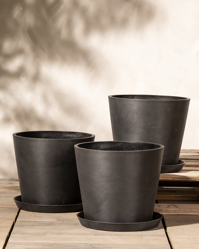Drei schwarze Pflanzkübel-Set Amsterdam (3x 30) in unterschiedlichen Größen werden auf einer Holzoberfläche präsentiert. Die Töpfe, jeweils mit passenden Untersetzern und integriertem Drainage-System, sind vor einem neutralen Hintergrund mit gesprenkelten Schatten positioniert und erzeugen eine ruhige, minimalistische Komposition.
