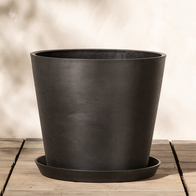 Ein schwarzer, zylindrischer Pflanzkübel aus Kunststoff (Amsterdam | 40 cm ⌀) mit passendem Untersetzer steht auf einer Holzfläche vor einer hellen, strukturierten Wand. Der Topf ist schlicht und modern im Design, mit klaren Linien und glatter Oberfläche. Sein integriertes Drainage-System sorgt für gesunde Pflanzen. Weiche Schatten werden auf Wand und Fläche geworfen.