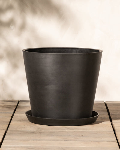 Ein großer, leerer schwarzer Blumentopf mit passendem Untersetzer steht auf einer Holzoberfläche. Dieser Kunststoff-Pflanzkübel (Amsterdam | 40 cm ⌀) hat eine konische Form und eine glatte, matte Oberfläche. Der Hintergrund ist eine schlichte, helle Wand mit subtilen Schatten, die das im Topf eingebaute Drainagesystem für optimale Pflanzengesundheit hervorhebt.