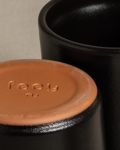 Nahaufnahme des Bodens zweier schwarzer Keramiktassen aus dem Keramik Baby-Topfset 'Sencillo' (9 Stk.). Die Tasse im Vordergrund steht umgedreht und gibt den Blick auf einen terrakottafarbenen Boden frei, auf dem der Markenname „feey“ eingeprägt ist. Die zweite Tasse steht aufrecht hinter der ersten Tasse und zeigt ihr schlichtes Design. Der Hintergrund ist neutral.
