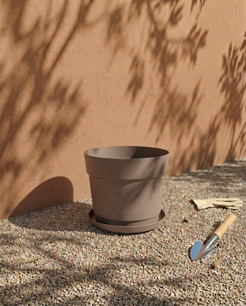 Ein großer leerer Kunststoff-Pflanzkübel (Bloom | 19 cm Ø) aus recyceltem Kunststoff sitzt auf Kies neben einer Stuckwand mit Blattschatten. Rechts neben dem UV-beständigen Topf liegen Gartenhandschuhe und eine kleine Handkelle.