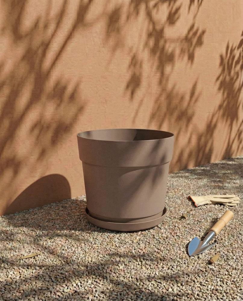 Ein großer, leerer Kunststoff Pflanzkübel (Bloom | 29 cm Ø), hergestellt aus recyceltem Kunststoff und UV-beständig, sitzt auf Kies neben einer Stuckwand mit Baumschatten. Gartenhandschuhe und eine kleine Kelle liegen daneben.