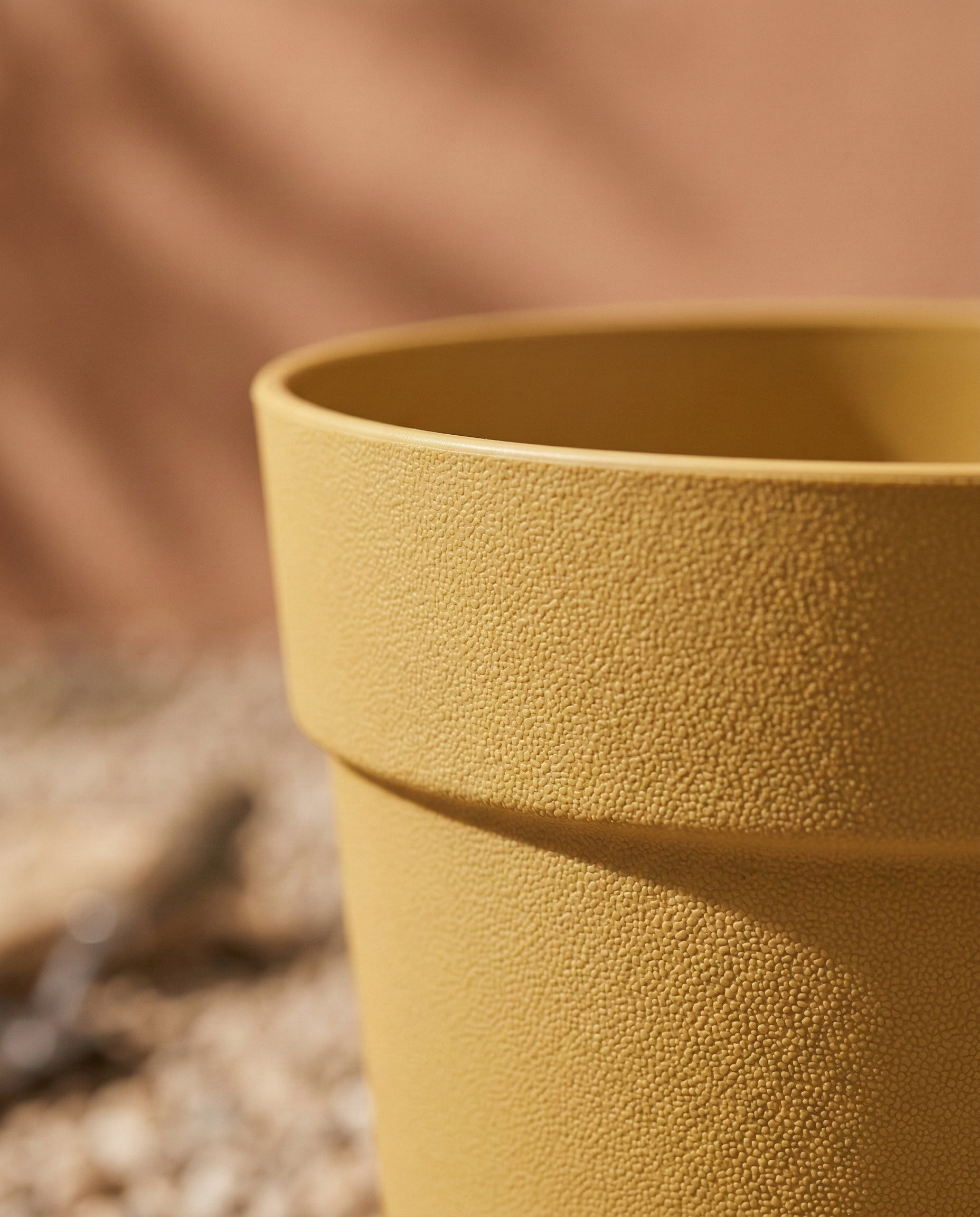 Nahaufnahme eines strukturierten gelben Kunststoff-Pflanzkübels (Bloom | 29 cm Ø) aus recyceltem Kunststoff, mit einem verschwommenen Hintergrund in warmen Braun- und Beigetönen und Kieselsteinen auf dem Boden.