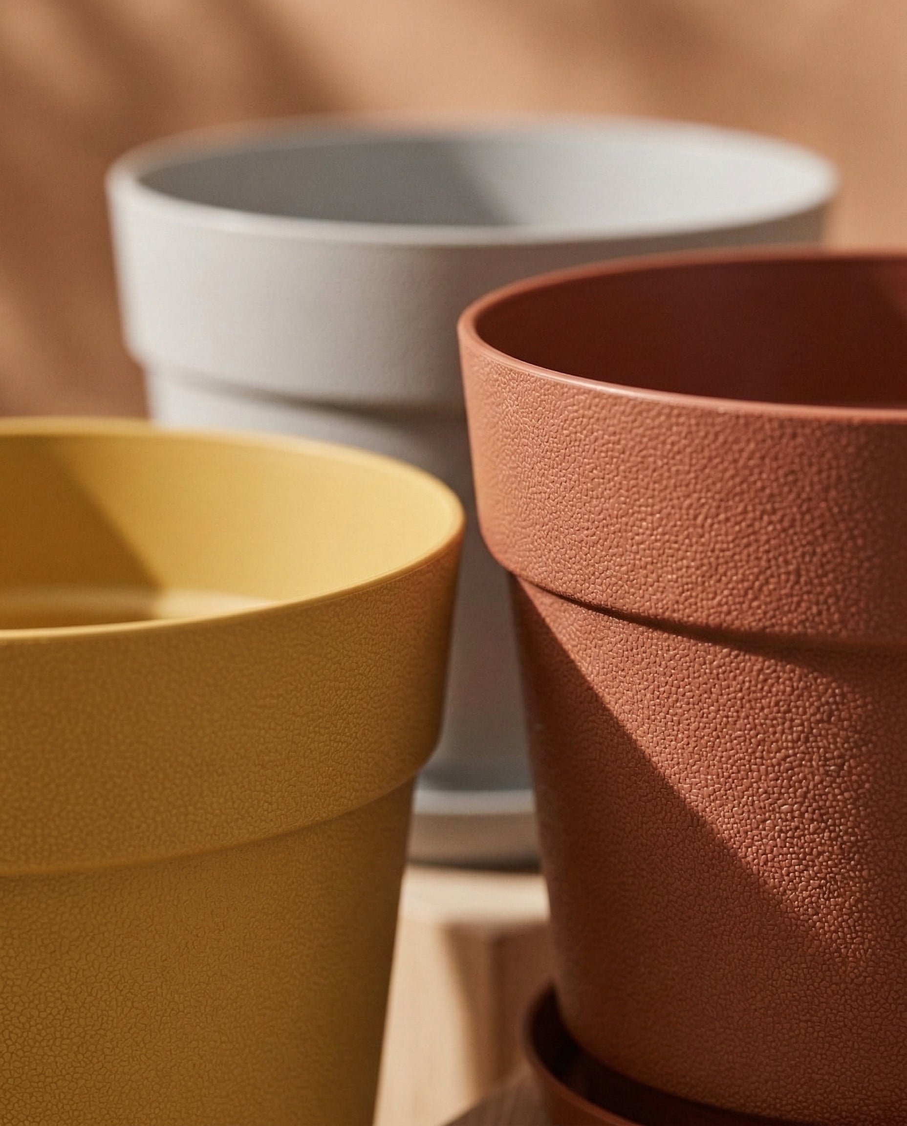 Drei leere Kunststoff-Pflanzkübel (Bloom | 29 cm Ø) in Gelb, Terrakotta und Hellgrau sind in Nahaufnahme mit strukturierten Oberflächen aus recyceltem Kunststoff zu sehen. Sanftes Sonnenlicht und ein verschwommener, warmtoniger Hintergrund ergänzen die Szene.