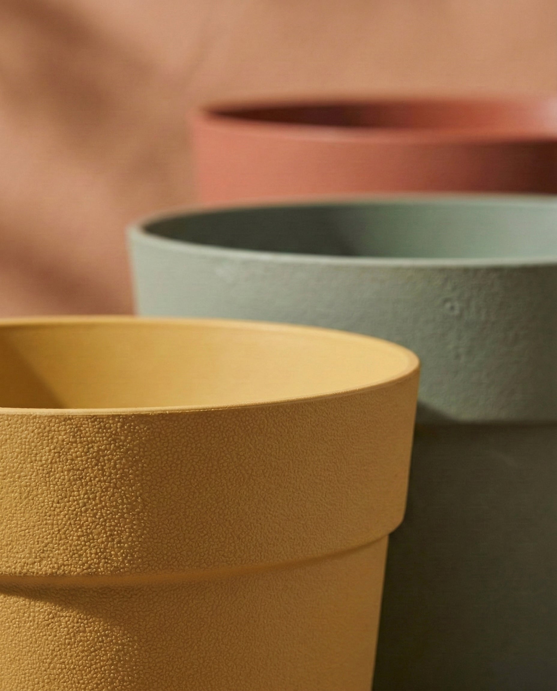 Drei Pflanzkübel aus dem 'Bloom'-Set (3× 29) aus recyceltem Kunststoff in Gelb, Grün und Terrakotta sind dicht nebeneinander auf einem weichen braunen Hintergrund angeordnet. Das schräge Foto hebt die Seiten und Ränder der strukturierten Töpfe hervor.