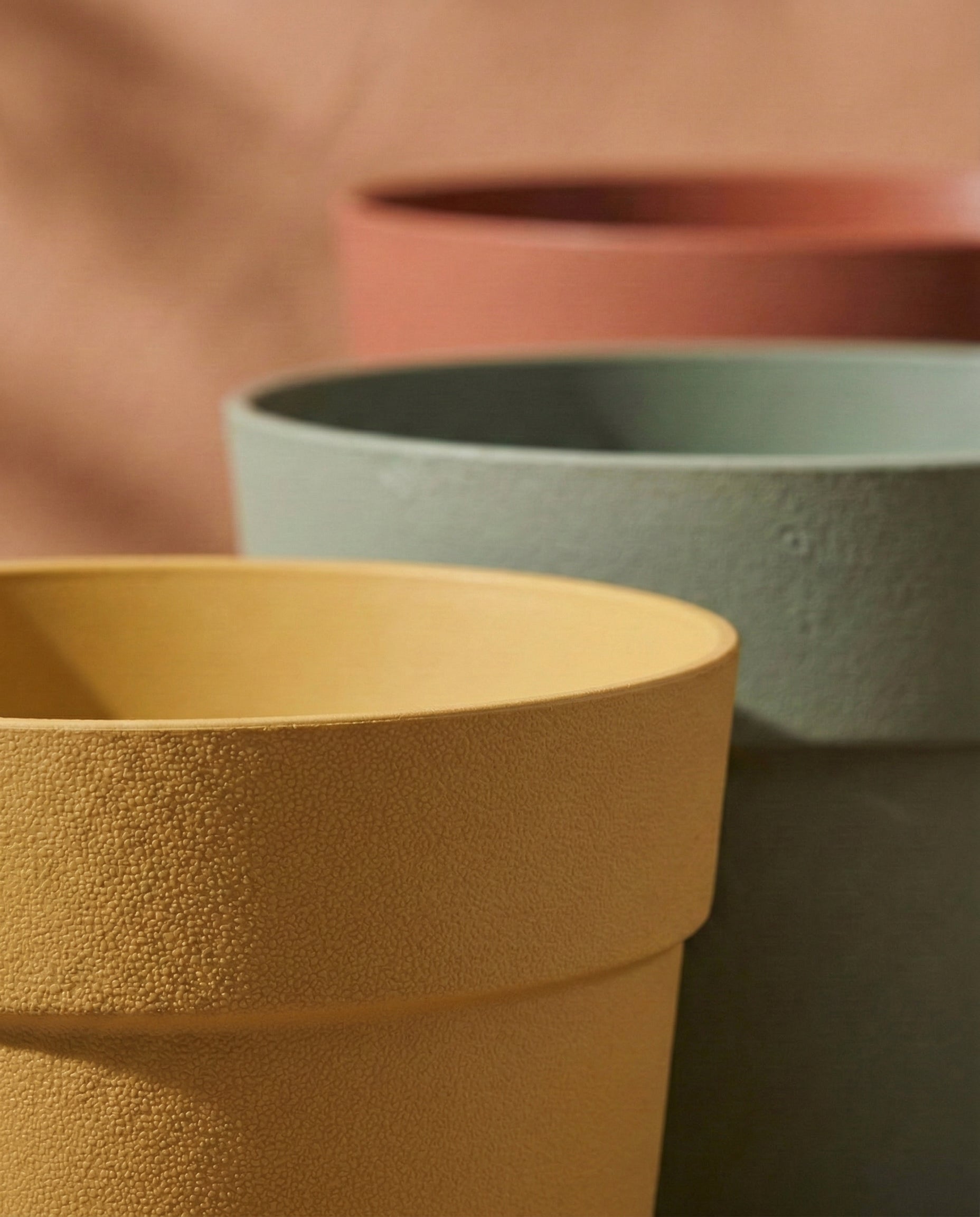 Drei Bloom Kunststoff Pflanzkübel (29 cm Ø) aus recyceltem Material in Gelb, Grün und Terrakotta, UV-beständig, sind vor neutralem Hintergrund eng gestapelt - sichtbar sind nur die oberen Ränder und Teile der Töpfe.