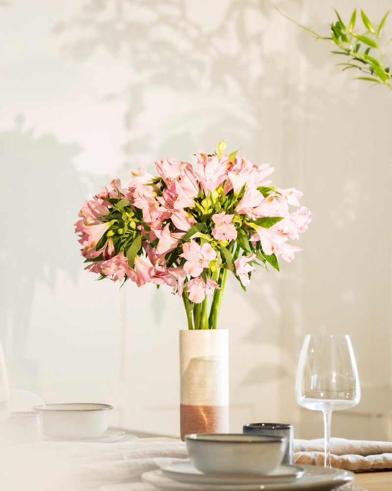 Eine fey Keramik-Vase (Variado | 21 cm) mit hellrosa Blüten steht auf einem Esstisch neben einem Weinglas und Keramikschalen, mit sanftem Sonnenlicht und Blätterschatten im Hintergrund.