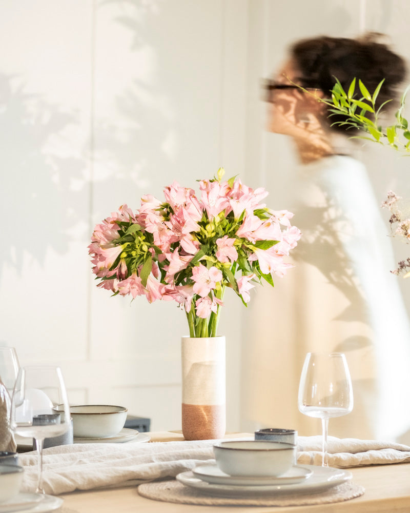 Eine unscharfe Person geht an einem gedeckten Esstisch vorbei, auf dem die Keramik-Vase (Variado | 21 cm) mit rosafarbenen Blumen in der Mitte steht, umgeben von Tellern, Schalen, Servietten und Weingläsern in sanften Neutraltönen.