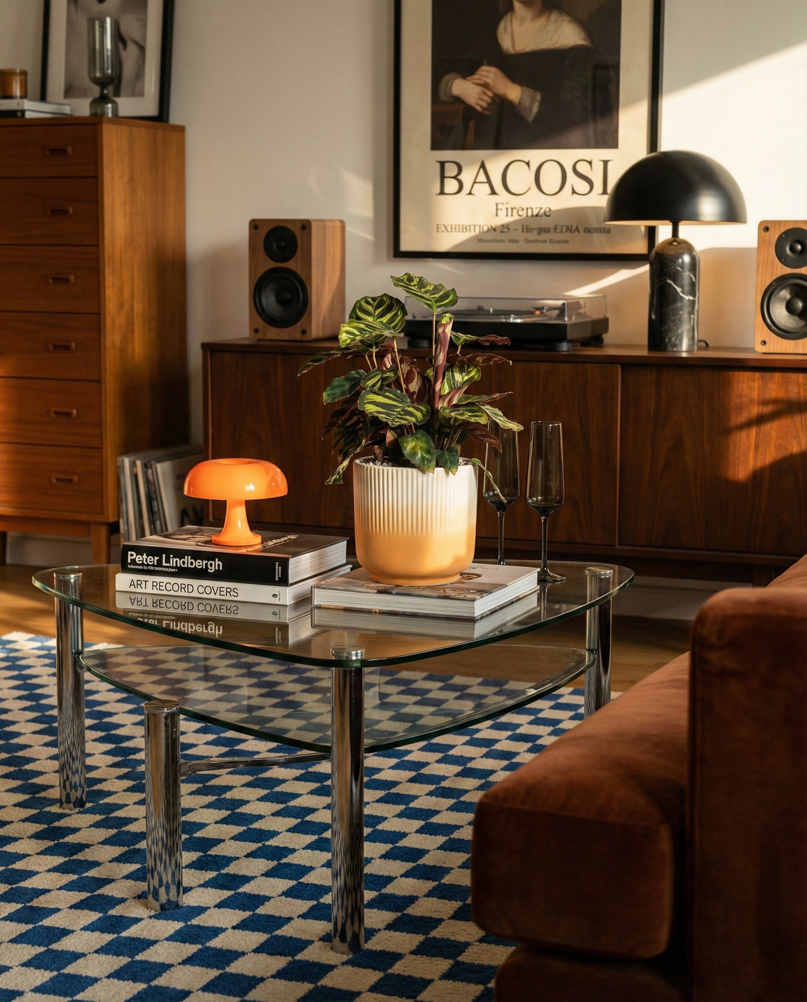 Ein modernes Wohnzimmer mit Dekor aus der Mitte des Jahrhunderts, mit einem gläsernen Couchtisch mit Büchern, einer orangefarbenen Lampe und der Kleinen Calathea makoyana - feey. Im Hintergrund stehen ein hölzernes Sideboard, Lautsprecher und gerahmte Kunstwerke.