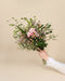Eine Hand in einem weißen Ärmel mit Spitzenmanschetten hält den Strauß Morgenlicht - von Hand gebunden mit gemischten Blumen, rosa Blüten, beerenartigen Akzenten und Grünzeug - und bringt die Wärme von Zuhause vor einem schlichten beigen Hintergrund.