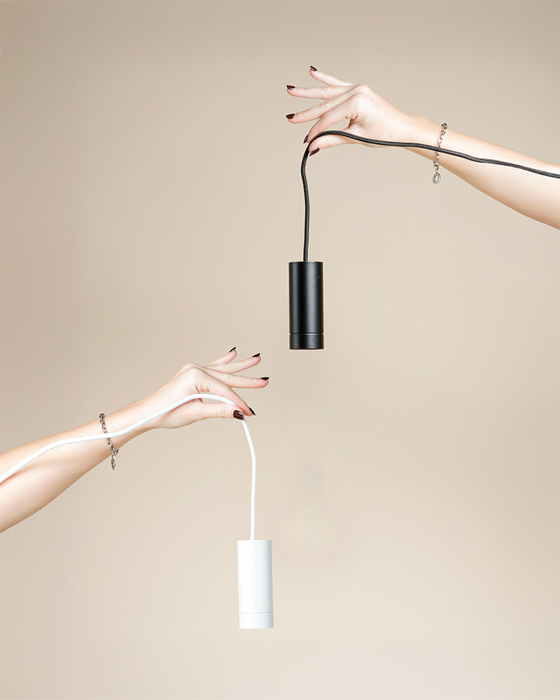 Zwei Hände mit manikürten Nägeln und silbernen Armbändern strecken sich vor einem beigen Hintergrund einander entgegen, jede hält eine Pendelleuchte 'Corda' - eine schwarz, eine weiß - beide zeigen das angebrachte Kabel.