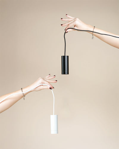 Zwei Hände mit manikürten Nägeln und silbernen Armbändern strecken sich vor einem beigen Hintergrund einander entgegen, jede hält eine Pendelleuchte 'Corda' - eine schwarz, eine weiß - beide zeigen das angebrachte Kabel.