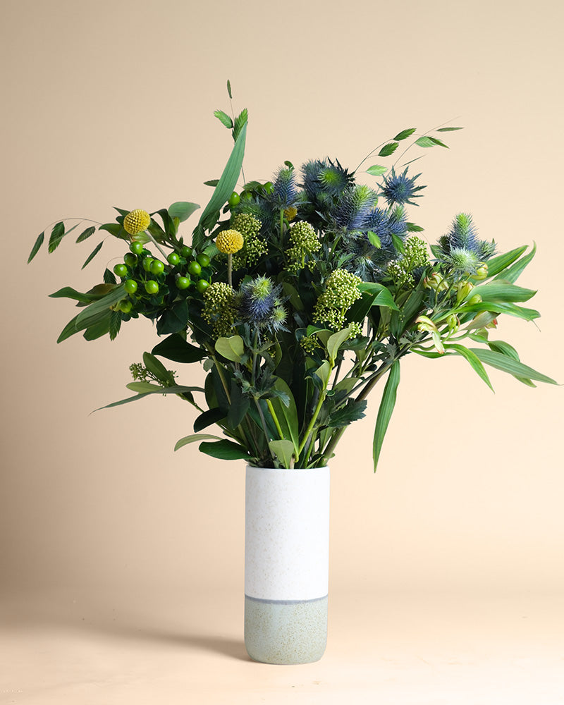 Die weiße Keramikvase "Erfolgsgeheimnis" zeigt grünes Laub, runde gelbe Bälle und stachelige blaue Distelblüten vor einem beigen Hintergrund und vermittelt ein Gefühl von Gelassenheit und positiver Energie.