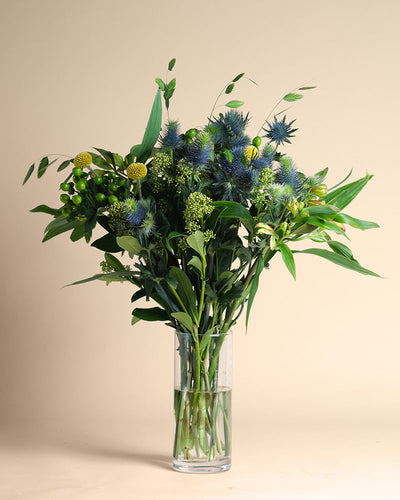 Die Vase Erfolgsgeheimnis zeigt ein klares Glasdesign, gefüllt mit grünen Blättern, gelben Bällen, blauen Disteln und strukturiertem Grün, das vor einem schlichten beigen Hintergrund positive Energie ausstrahlt.