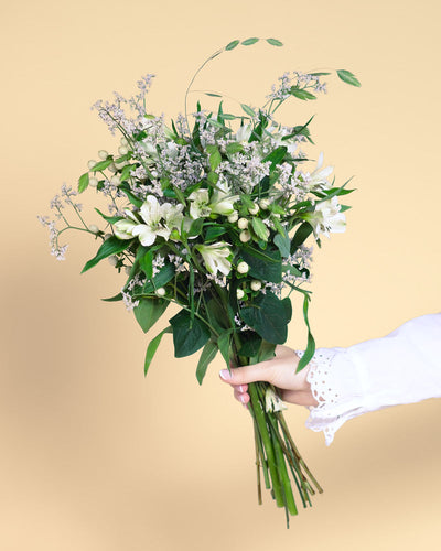 Eine Hand in einem weißen Rüschenärmel hält Luftkuss - einen Strauß aus grünem Laub, weißen Lilien, kleinen weißen Beeren und zarten Blüten - und flüstert Liebe vor einem hellbeigen Hintergrund.