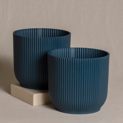 Zwei marineblaue gerippte Topfsets (2 x 16) unterschiedlicher Größe stehen auf einer beigen Oberfläche. Der größere Topf steht auf einer kleinen hellbraunen Box, während der kleinere Topf direkt auf der Oberfläche steht. Der Hintergrund ist ein schlichter beiger Stoff.