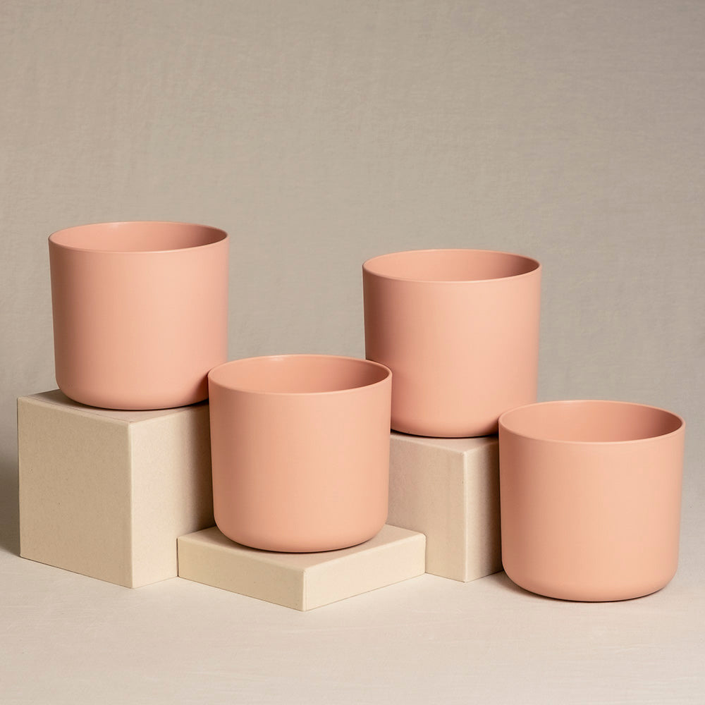 Vier pfirsichfarbene Keramik-Topfsets (4 x 14) in verschiedenen Größen sind auf und um beige würfelförmige Ständer angeordnet, die vor einem neutralen, strukturierten Hintergrund platziert sind. Die Töpfe haben ein minimalistisches Design und eine glatte Oberfläche, perfekt für die Präsentation von Zimmerpflanzen.