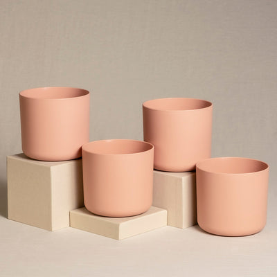 Vier pfirsichfarbene Keramik-Topfsets (4 x 14) in verschiedenen Größen sind auf und um beige würfelförmige Ständer angeordnet, die vor einem neutralen, strukturierten Hintergrund platziert sind. Die Töpfe haben ein minimalistisches Design und eine glatte Oberfläche, perfekt für die Präsentation von Zimmerpflanzen.