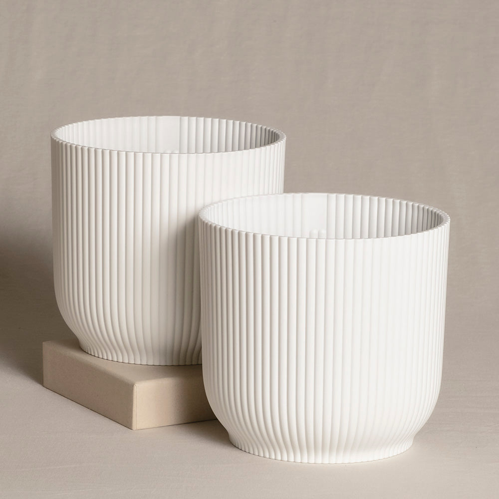 Zwei weiße, zylindrische dekorative Topfsets (2 x 16) mit vertikalem Rippenmuster. Ein Topf ist etwas größer, beide stehen auf einer neutral getönten Oberfläche, wobei der größere teilweise auf einem beigen rechteckigen Block steht. Der Hintergrund ist ein weicher, beiger Stoff.