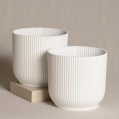 Zwei weiße, zylindrische dekorative Topfsets (2 x 16) mit vertikalem Rippenmuster. Ein Topf ist etwas größer, beide stehen auf einer neutral getönten Oberfläche, wobei der größere teilweise auf einem beigen rechteckigen Block steht. Der Hintergrund ist ein weicher, beiger Stoff.