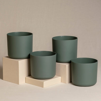 Vier dunkelgrüne Keramiktopfsets (4 x 14) mit glatter, matter Oberfläche sind auf und um drei beigefarbene quadratische Sockel angeordnet. Der Hintergrund ist neutral, mit einem sanften Beigeton, der das minimalistische und elegante Design der Pflanzgefäße ergänzt.