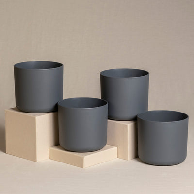 Vier dunkelgraue zylindrische Topfsets (4 x 14) sind auf und um drei helle rechteckige Blöcke vor einem beigen Hintergrund platziert. Die Pflanzgefäße haben ein einfaches, modernes Design und sind gleichmäßig verteilt.