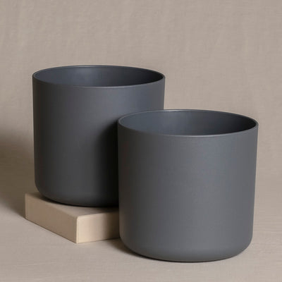 Zwei dunkelgraue zylindrische Topfsets (2 x 16) sind vor einem beigen Hintergrund zu sehen. Der größere Topf steht etwas links hinter dem kleineren, der auf einem kleinen beigen Block steht. Das Design ist schlicht und modern, mit einem matten Finish.