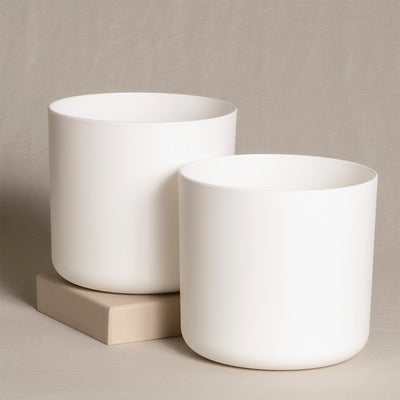 Zwei weiße, zylindrische Topfsets (2 x 16) stehen auf einer hellbeigen Oberfläche. Ein Topf ist etwas größer als der andere. Der größere Topf steht direkt auf der Oberfläche, während der kleinere auf einem kleinen, beigen Block steht. Der Hintergrund ist neutral.
