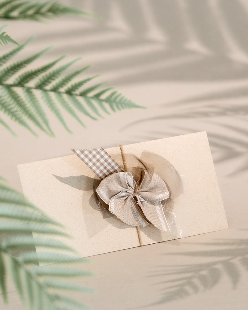 Ein beiger Gutschein mit dekorativer Schleife und kariertem Band liegt auf einer hellen Unterlage. Farnblätter werfen im Vordergrund sanfte Schatten und verleihen der eleganten Präsentation eine natürliche Note.