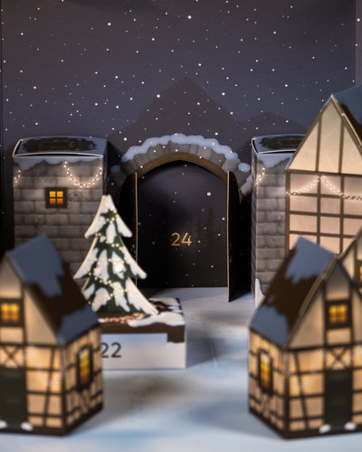 Eine bezaubernde Adventskalender-Szene mit einem verschneiten Winterdorf. Der Pflanzen-Adventskalender zeigt beleuchtete Papierhäuser und einen wunderschön geschmückten Baum im Vordergrund. In der Mitte ist eine Tür mit der Aufschrift „24“ von schneebedeckten Gebäuden unter einem sternenklaren Nachthimmel umgeben.