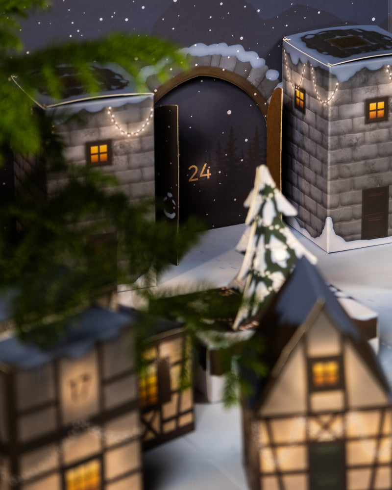Eine festliche Szene präsentiert den entzückenden Pflanzen-Adventskalender mit schneebedeckten Häusern und einem geschmückten Tannenbaum. Ein Adventskalender-Fenster mit der Aufschrift „24“ ist geöffnet und gibt den Blick auf eine verschneite Nacht frei, die mit funkelnden Sternen und festlichen Lichtern geschmückt ist.