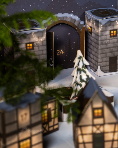 Eine festliche Szene präsentiert den entzückenden Pflanzen-Adventskalender mit schneebedeckten Häusern und einem geschmückten Tannenbaum. Ein Adventskalender-Fenster mit der Aufschrift „24“ ist geöffnet und gibt den Blick auf eine verschneite Nacht frei, die mit funkelnden Sternen und festlichen Lichtern geschmückt ist.