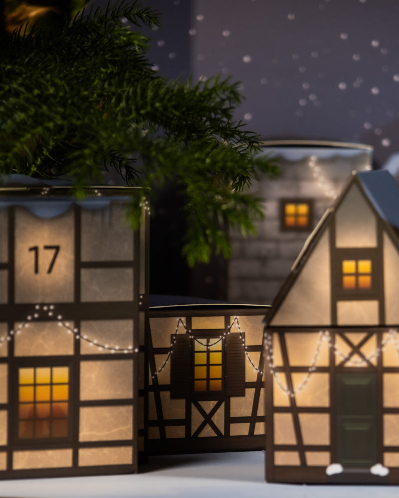 Eine festliche Darstellung des Pflanzen-Adventskalenders besteht aus beleuchteten Miniatur-Papierhäusern mit hübschen Holzrahmen. Ihre winzigen Fenster strahlen ein warmes Licht aus, das durch Lichterketten an der Außenseite ergänzt wird. Darüber hängt ein Tannenzweig vor dem Hintergrund eines dunklen Sternenhimmels und schafft eine einladende Weihnachtsatmosphäre.