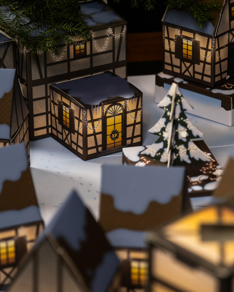 Eine gemütliche Winterdorfszene erinnert an den Charme des Pflanzen-Adventskalenders und zeigt Papierhäuser mit leuchtenden Fenstern und schneebedeckten Dächern. Abgerundet wird die Winterdorf-Ausstellung durch einen geschmückten Weihnachtsbaum vor weißem Hintergrund, der eine festliche und warme Weihnachtsstimmung hervorruft.