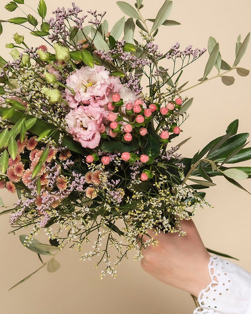 Eine Hand in einem weißen Spitzenärmel hält den Morgenlicht-Strauß aus gemischten Blumen - mit rosa Blüten, kleinen Beeren und Grünzeug - vor einem beigen Hintergrund.