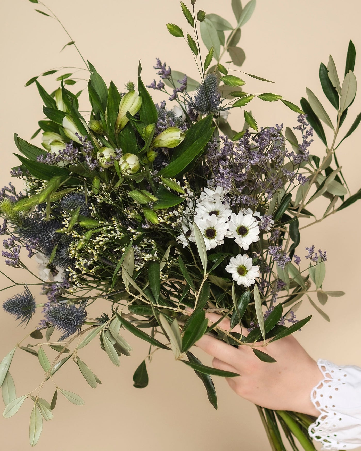 Eine Hand hält den zarten Blumenstrauß "Nebelmeer" aus weißen Gänseblümchen, grünen Blättern, lilafarbenen distelartigen Blüten und Lavendel im Morgentau vor beigem Hintergrund.