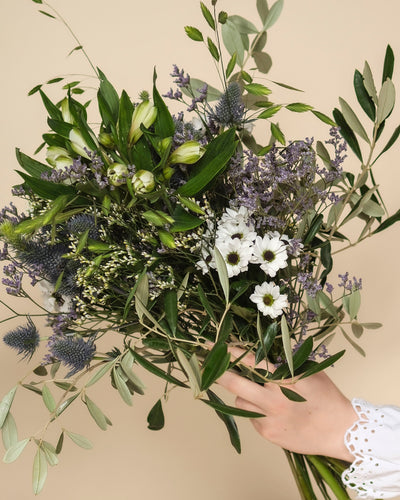 Eine Hand hält den zarten Blumenstrauß "Nebelmeer" aus weißen Gänseblümchen, grünen Blättern, lilafarbenen distelartigen Blüten und Lavendel im Morgentau vor beigem Hintergrund.