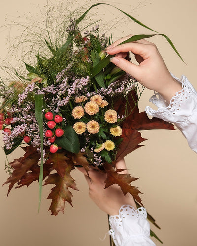 Eine Person in einer weißen Bluse mit Ösenmanschetten arrangiert den Herbstfreude-Strauß - gemischte Blumen, rote Beeren und herbstliches Laub in warmen Farben - vor einem beigen Hintergrund, so dass ein fröhliches Farbenspiel in ihren Händen entsteht.
