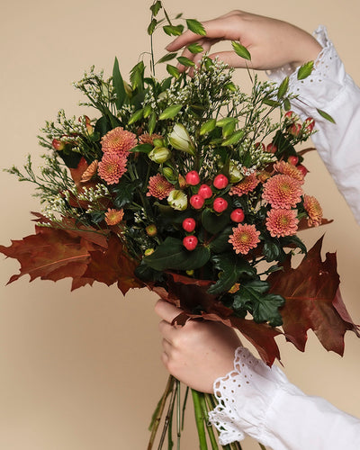 Eine Person in einer weißen, langärmeligen Bluse mit Spitzenmanschetten hält den Herbstfreude-Strauß - rosafarbene Chrysanthemen, rote Beeren, grüne Knospen, weiße Blüten und braune Blätter - und schafft damit ein lebendiges Farbspiel vor einem beigen Hintergrund.