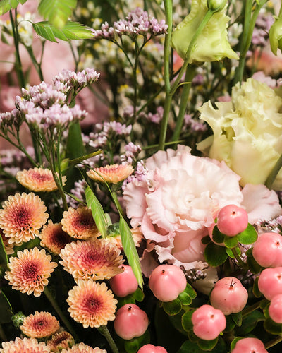 Eine Nahaufnahme des farbenfrohen Blumenarrangements Morgenlicht, von Hand gebunden, mit rosafarbenen Nelken, blassgelben Lisianthus, pfirsichfarbenen Chrysanthemen, lila Statice und rosafarbenen Beerenbüscheln - perfekt, um Ihr Zuhause zu verschönern.