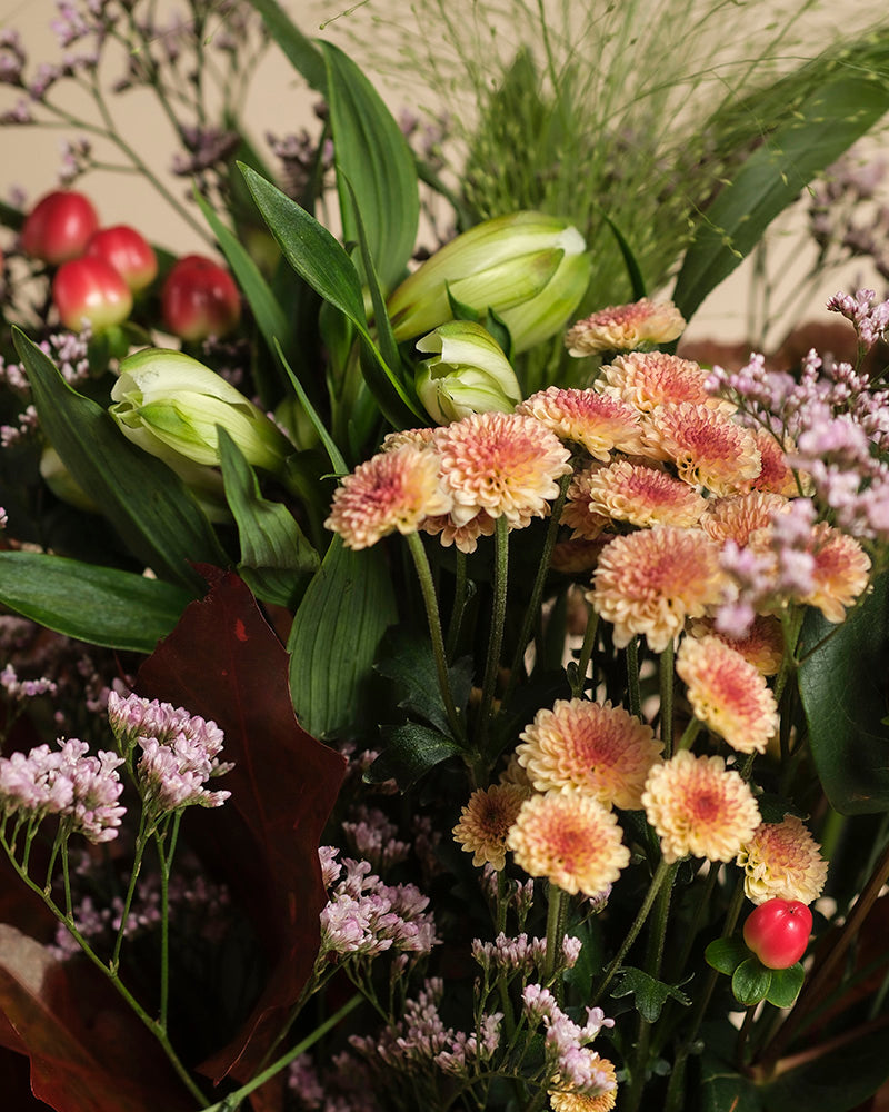 Eine Nahaufnahme von Herbstfreude zeigt pfirsichfarbene Chrysanthemen, hellviolette Statice, geschlossene weiße Lilien, grüne Blätter und rote Hypericum-Beeren in warmen Tönen für ein fröhliches Farbenspiel.