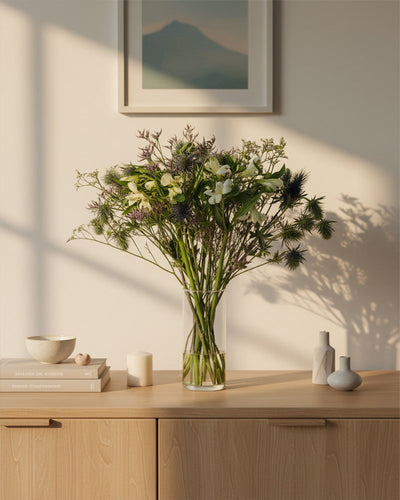 In einer klaren Glasvase steht der grün-weiße Wanderlust-Strauß und bringt Bergluft-Stimmung in einen hellen Holzschrank. In der Nähe liegen Bücher, Kerzen und kleine Vasen, während das Sonnenlicht sanfte Schatten unter die gerahmte Wandkunst wirft.
